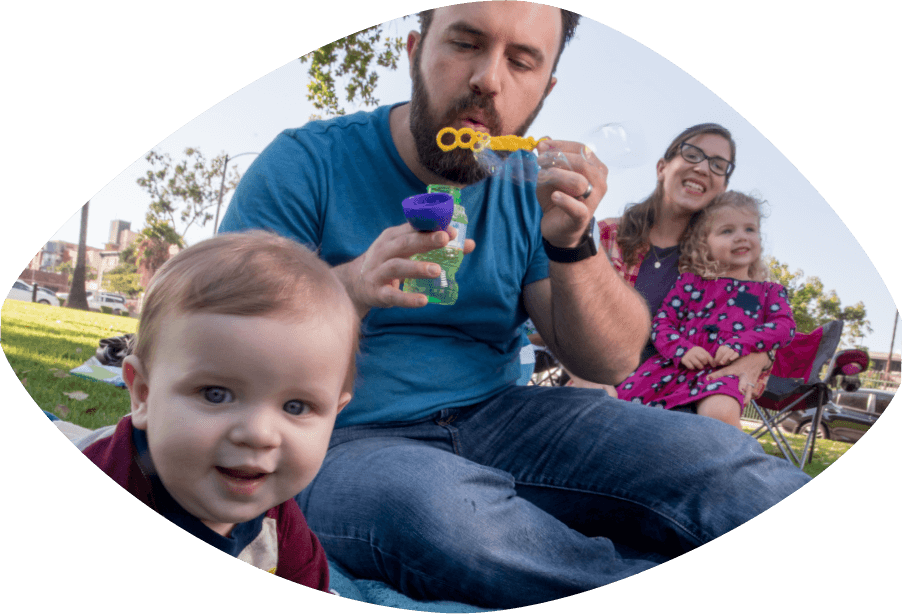 Papá y mamá están sentados en un parque mientras que papá sopla burbujas para su niñita y niñito chiquitos.