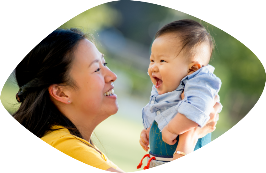 Una sonriente mamá asiática carga a su niño chiquito frente a sí, mientras que el niñito se rí y mira a su mamá.