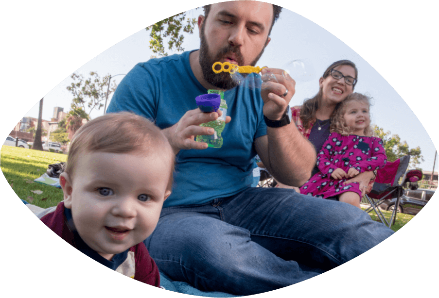 Father and mother sitting are in a park while the father blows bubbles for their small boy and girl