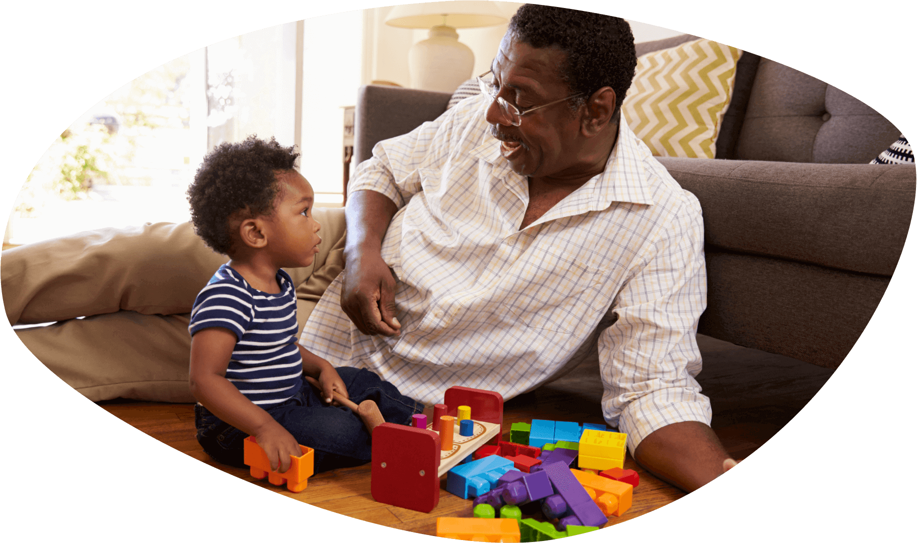 An African American toddler is looking at his laughing father as they sit on the floor playing with giant legos